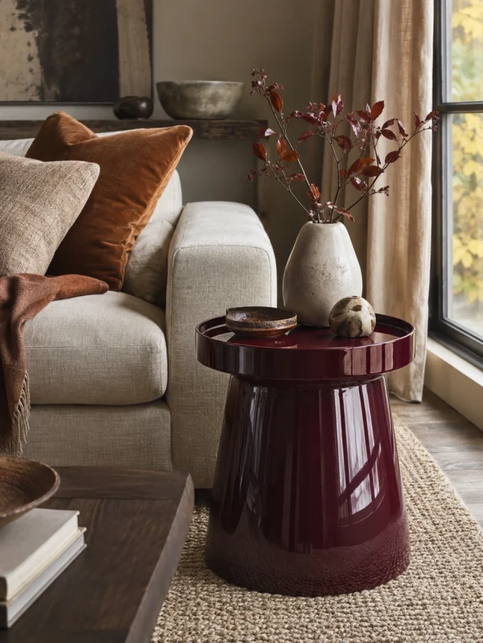 A glossy, dark red side table with a vase and small objects on top, next to a beige couch with brown pillows.