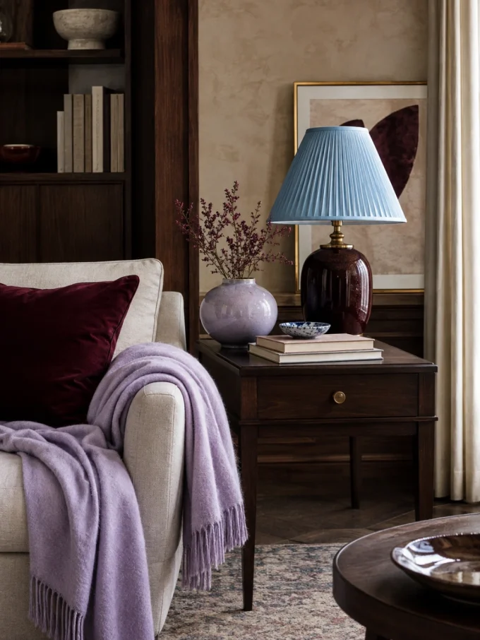 A cream armchair with a lilac throw blanket and maroon pillow beside a dark wood side table with a blue lamp, lilac vase, and books.